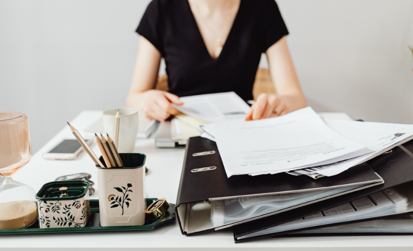 SachbezugSchreibtisch mit vielen Aktenordnern und Papierstapeln, die eine Person im Hintergrund bearbeitet; Symbolbild für bürokratischen Aufwand bei Sachbezügen.